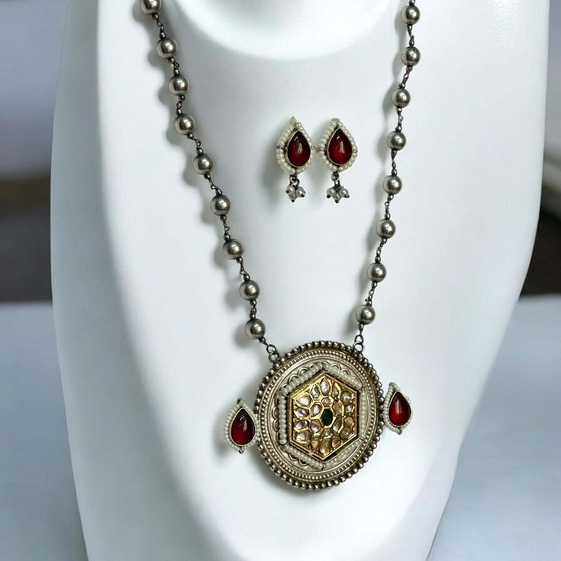 Dual tone Apala-style big-sized pendant set featuring a gold and silver finish with Kundan work, paired with red and green gemstone accents, displayed on a white jewellery display stand.