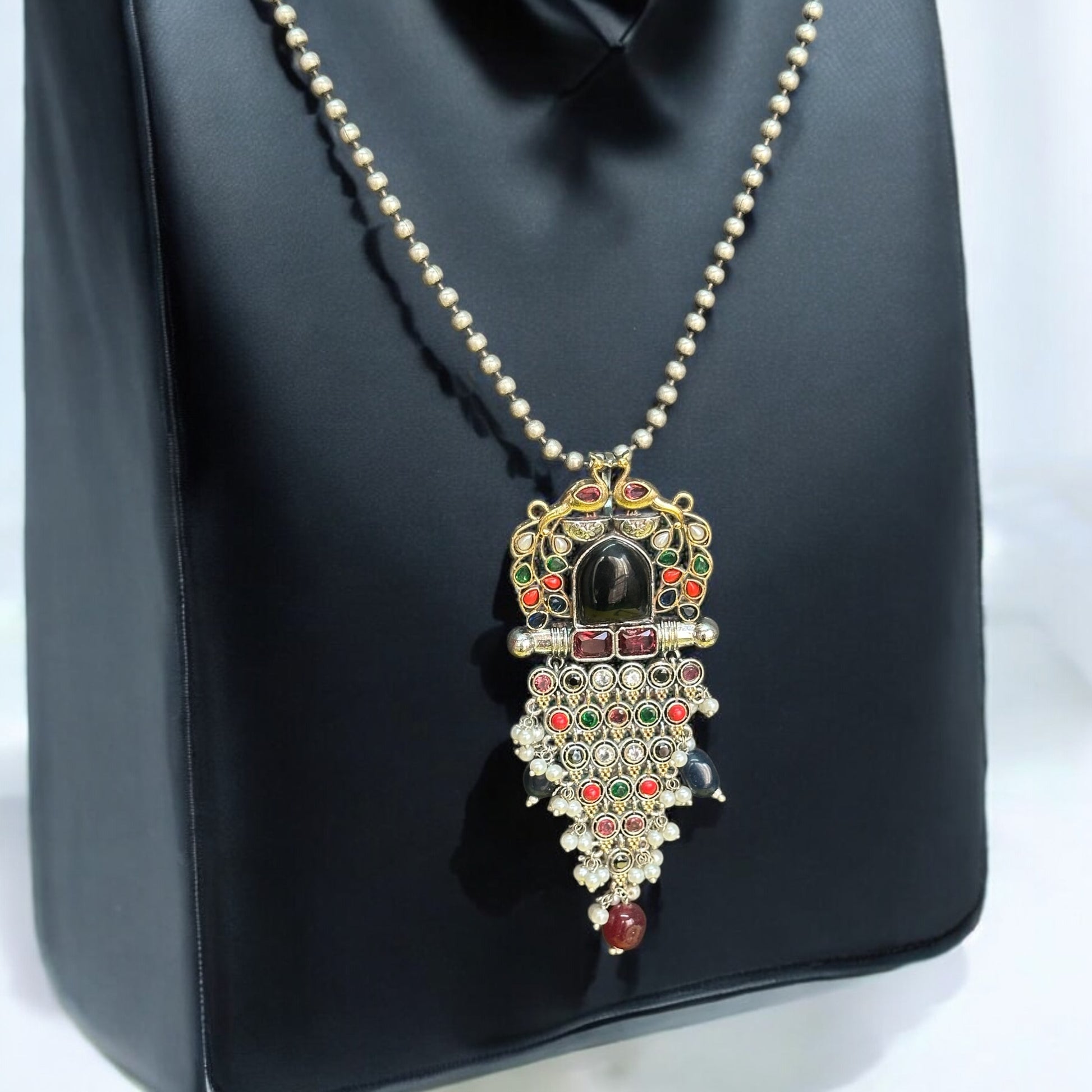 Long silver dual-tone peacock pendant set with intricate detailing, adorned with multicolor stones, pearls, and a matching pair of earrings, displayed against a black jewellery display stand.