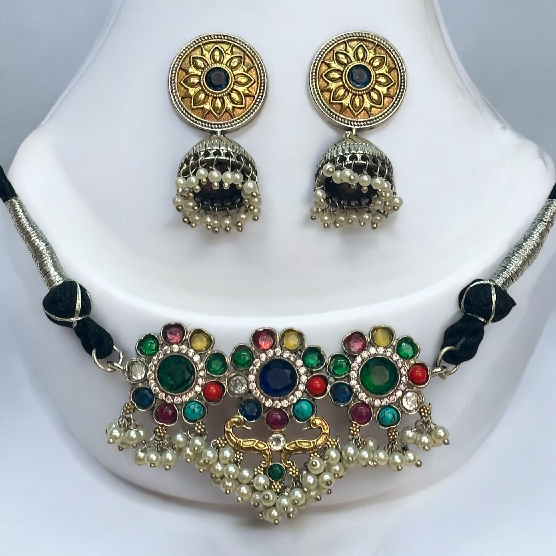 Minimal silver dual-tone choker necklace adorned with multicolor gemstones and pearl detailing, paired with intricate jhumki earrings, displayed on a white jewellery display stand in a light background.
