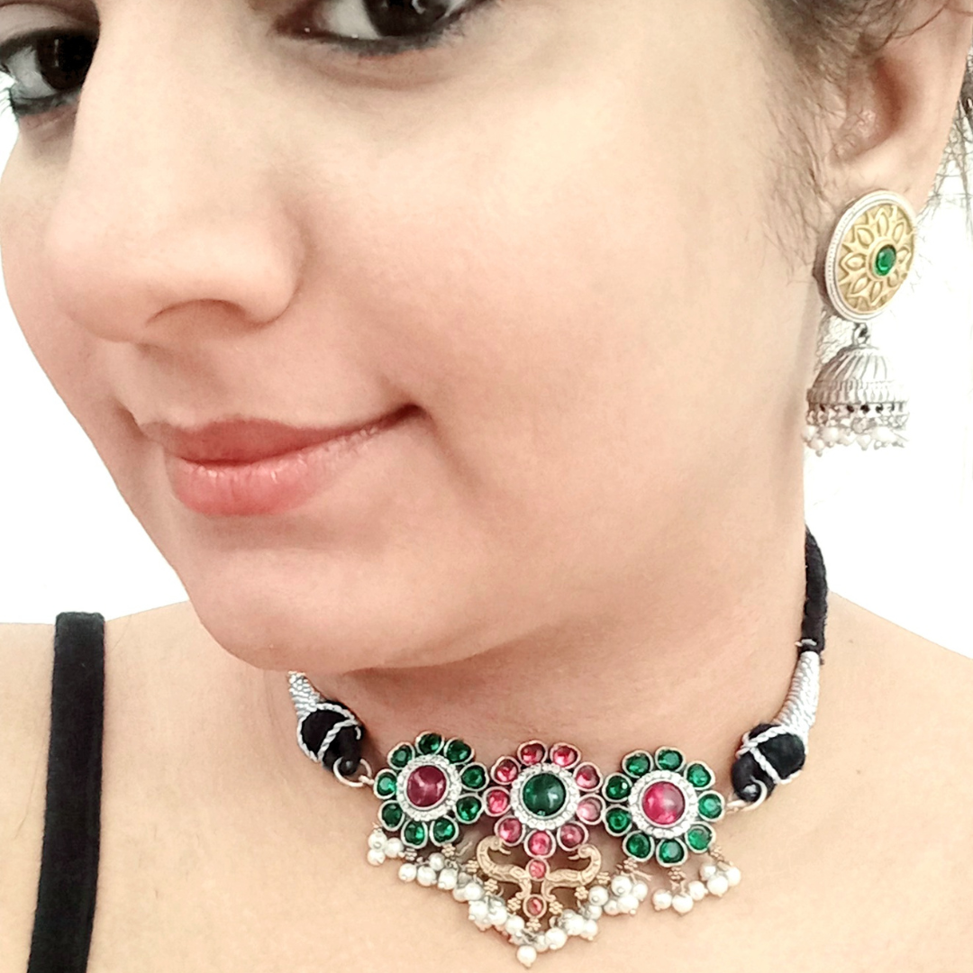 A model wearing a Minimal silver dual-tone choker featuring intricate floral motifs with green and pink gemstones, accented with pearls. Paired with gold-toned round jhumka earrings with detailed patterns, displayed on a white background.