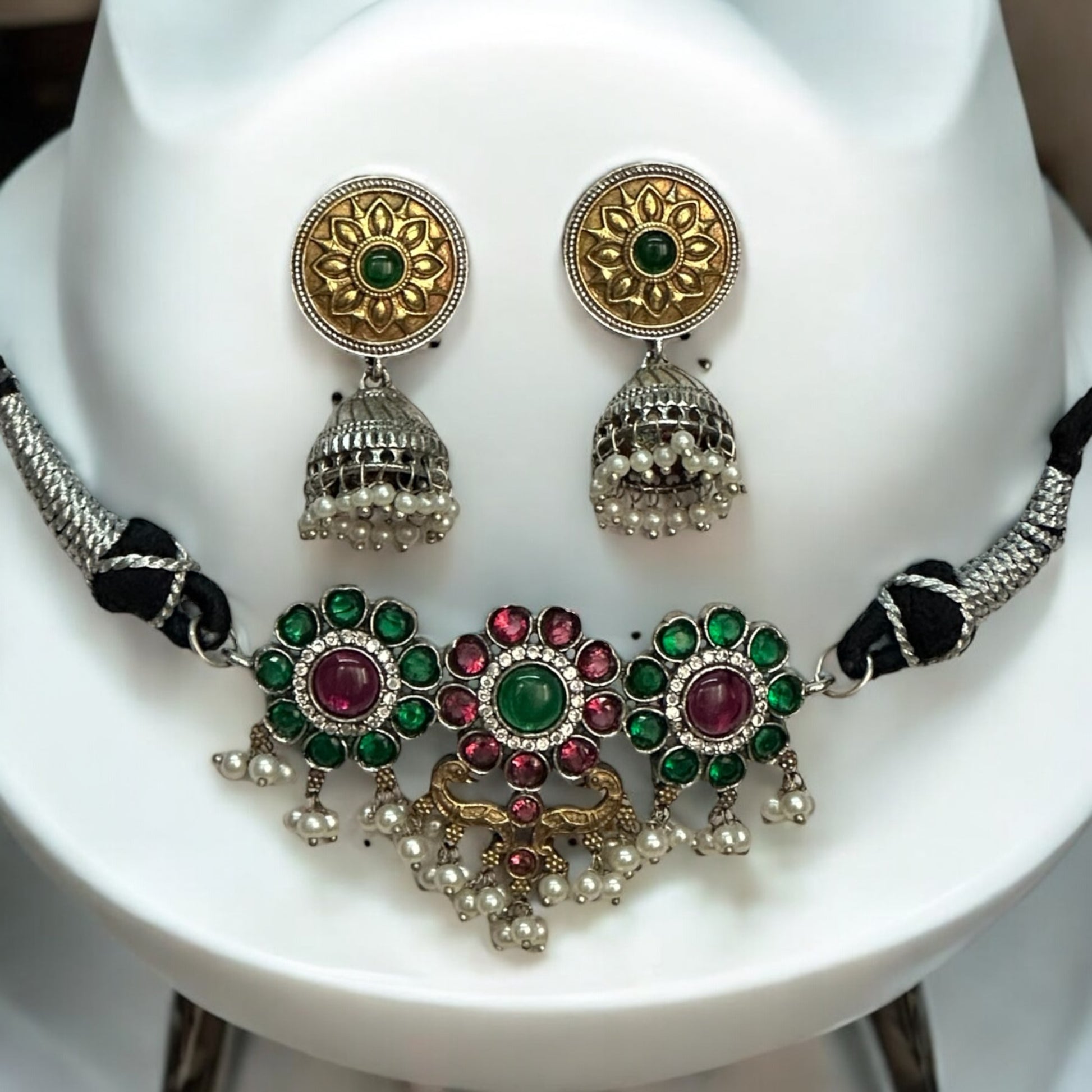 Minimal silver dual-tone choker featuring intricate floral motifs with green and pink gemstones, accented with pearls. Paired with gold-toned round jhumka earrings with detailed patterns, displayed on a white background.