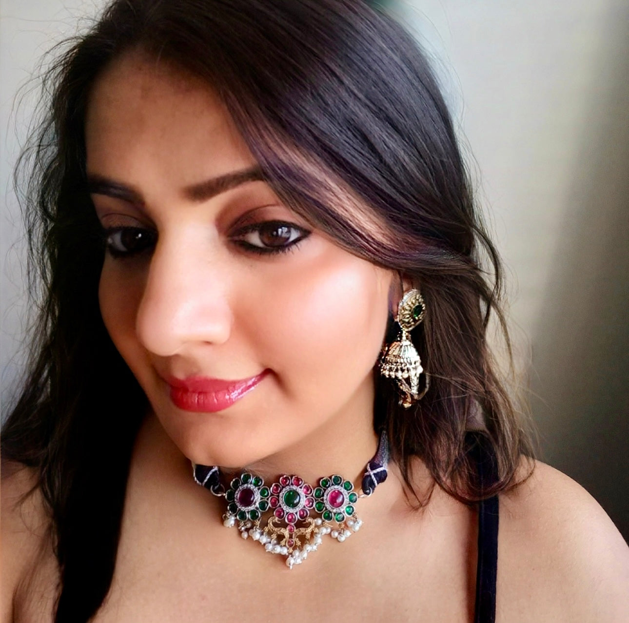A model displaying Minimal silver dual-tone choker featuring intricate floral motifs with green and pink gemstones, accented with pearls. Paired with gold-toned round jhumka earrings with detailed patterns, to sell online in london via bella n diva.