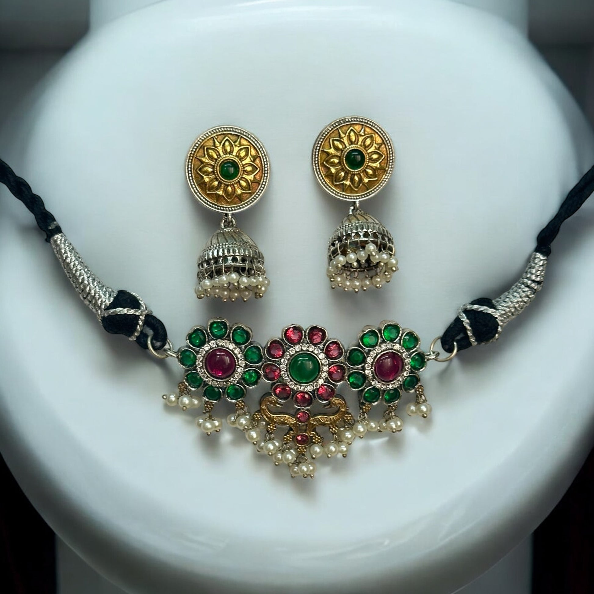 Minimal silver dual-tone choker featuring intricate floral motifs with green and pink gemstones, accented with pearls. Paired with gold-toned round jhumka earrings with detailed patterns, displayed on a white jewellery display stand.