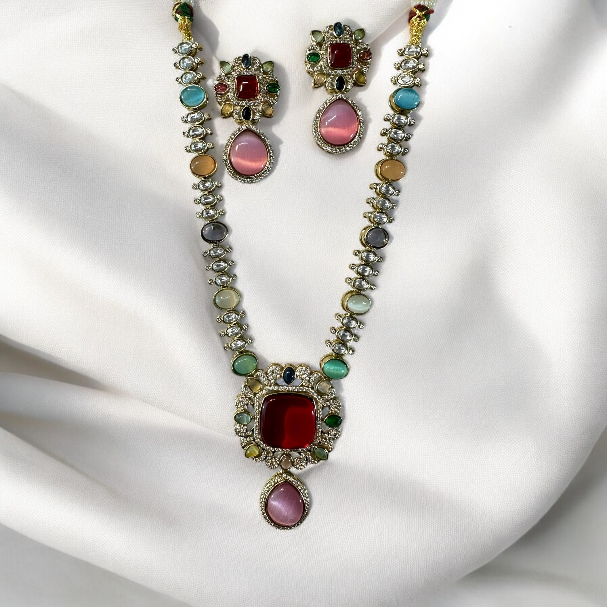 Moissanite Premium Multicolor Kundan Necklace Set WITH EARRINGS PLACED ON A WHITE COLORED FABRIC. iT HAS RED STONES BOTH IN NECKLACE AND EARRINGS AND ON THE NECKLACE STRING, THERE ARE MULTICOLORED STONES WITH SHINY CRYSTALS EMBEDED.