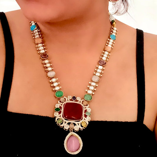 A model wearing a Moissanite Premium Multicolor Kundan Necklace Set WITH EARRINGS PLACED ON A WHITE COLORED FABRIC. iT HAS RED STONES BOTH IN NECKLACE AND EARRINGS AND ON THE NECKLACE STRING, THERE ARE MULTICOLORED STONES WITH SHINY CRYSTALS EMBEDED.