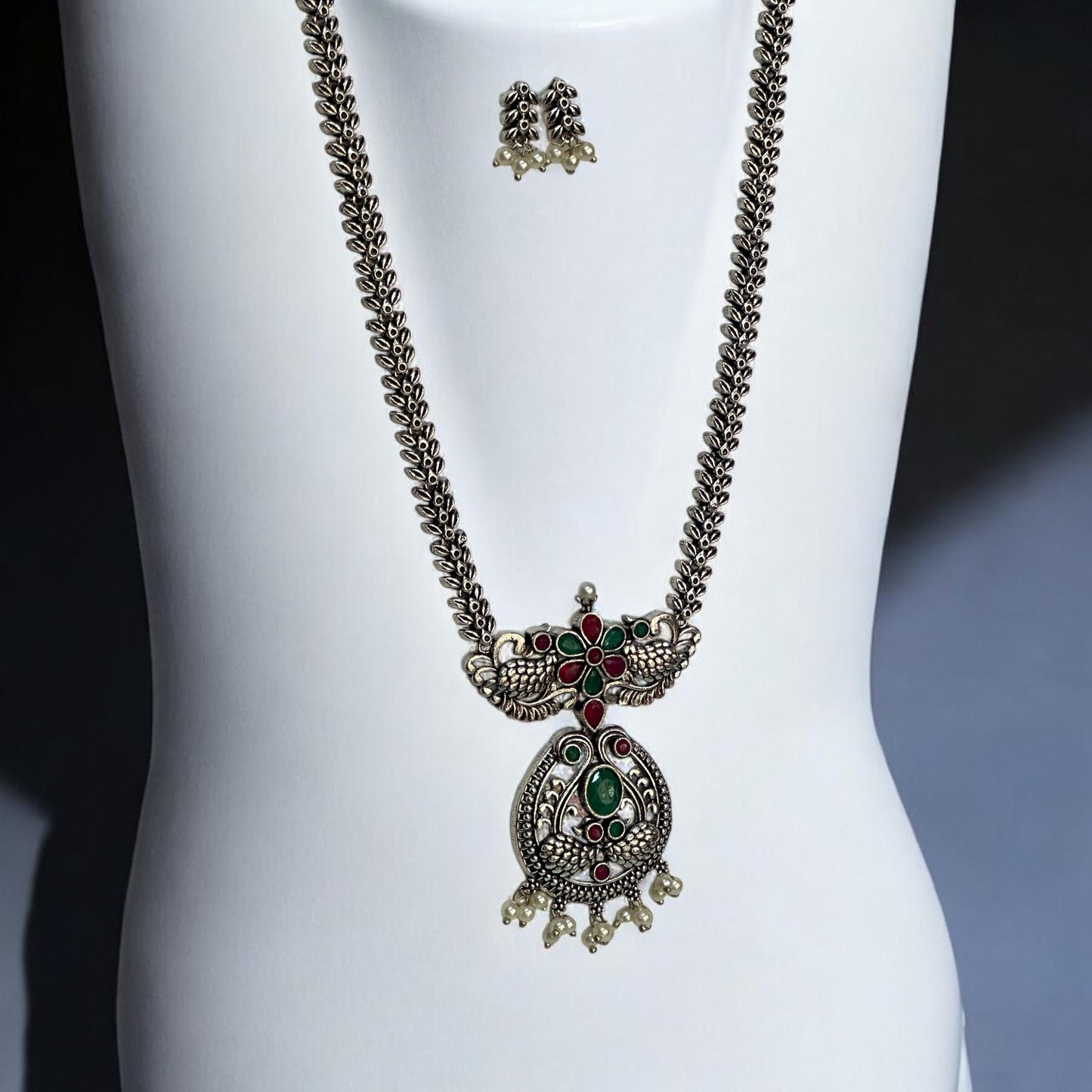 Side view of Oxidised long necklace set featuring ruby-red and emerald-green stones with a peacock-inspired pendant design, complemented by matching earrings with pearl drops, displayed on a white mannequin.