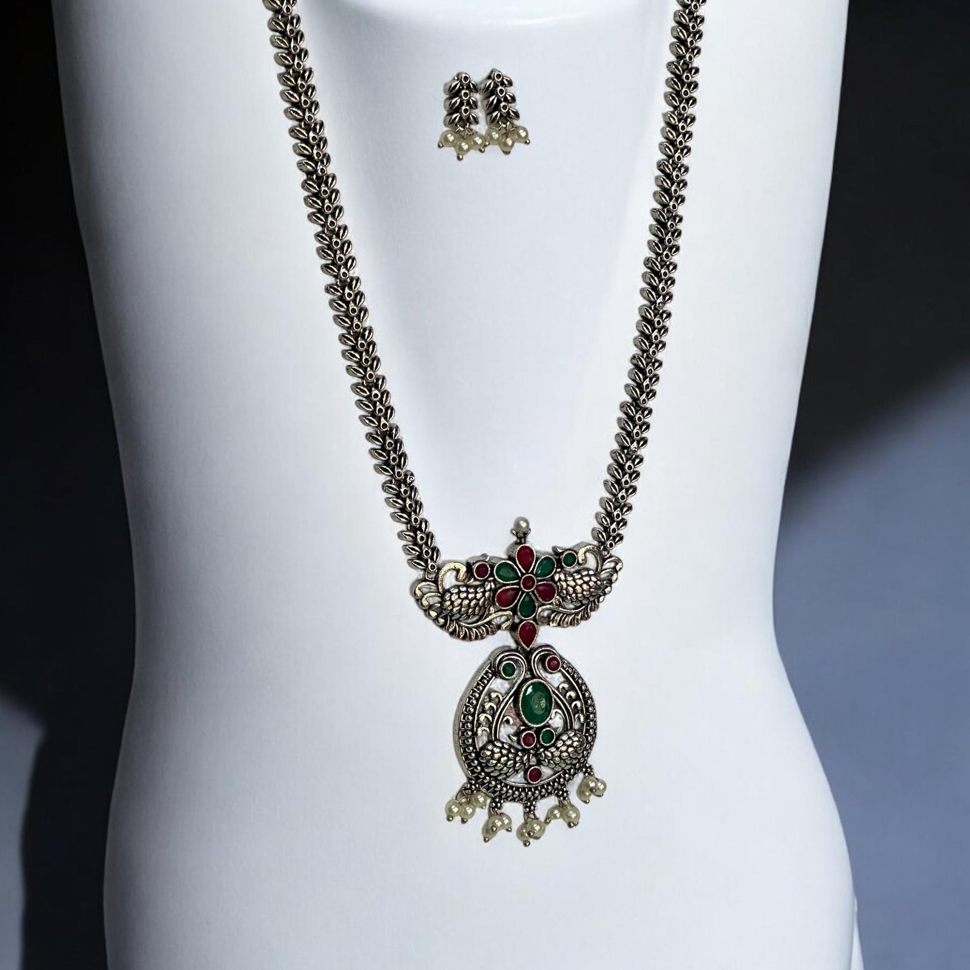 Side view of Oxidised long necklace set featuring ruby-red and emerald-green stones with a peacock-inspired pendant design, complemented by matching earrings with pearl drops, displayed on a white mannequin.