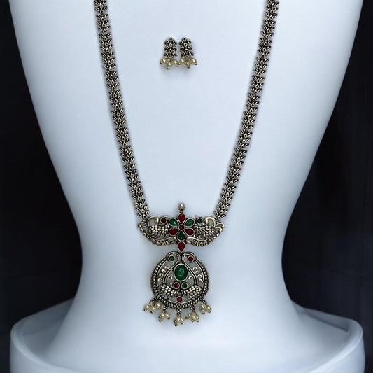Oxidised long necklace set featuring ruby-red and emerald-green stones with a peacock-inspired pendant design, complemented by matching earrings with pearl drops, displayed on a white mannequin.