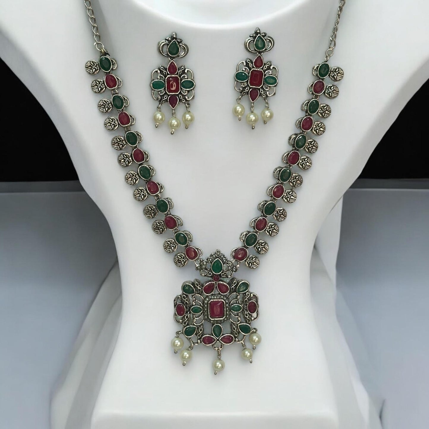 Oxidised necklace set featuring ruby-red and emerald-green stones with pearl drops, paired with matching earrings, displayed on a white mannequin against a soft background.