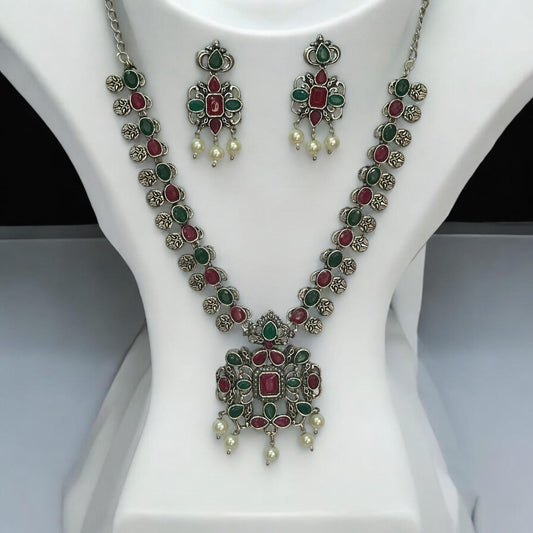 Oxidised necklace set featuring ruby-red and emerald-green stones with pearl drops, paired with matching earrings, displayed on a white mannequin against a soft background.