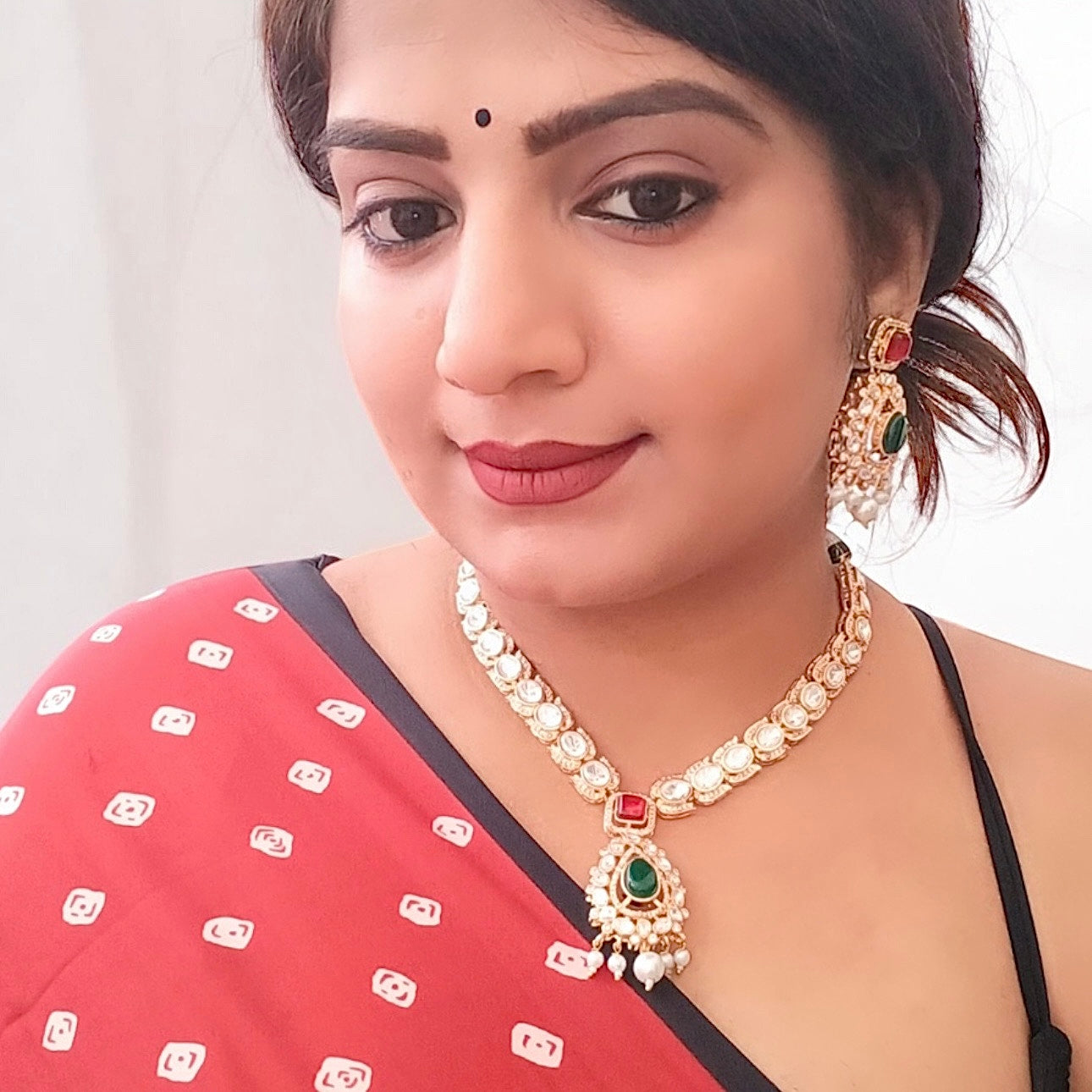 A model displaying A luxurious Moissanite necklace set with green and red stones, featuring intricate silver foil back finishing, Kundan-style settings, and delicate pearl droplets.