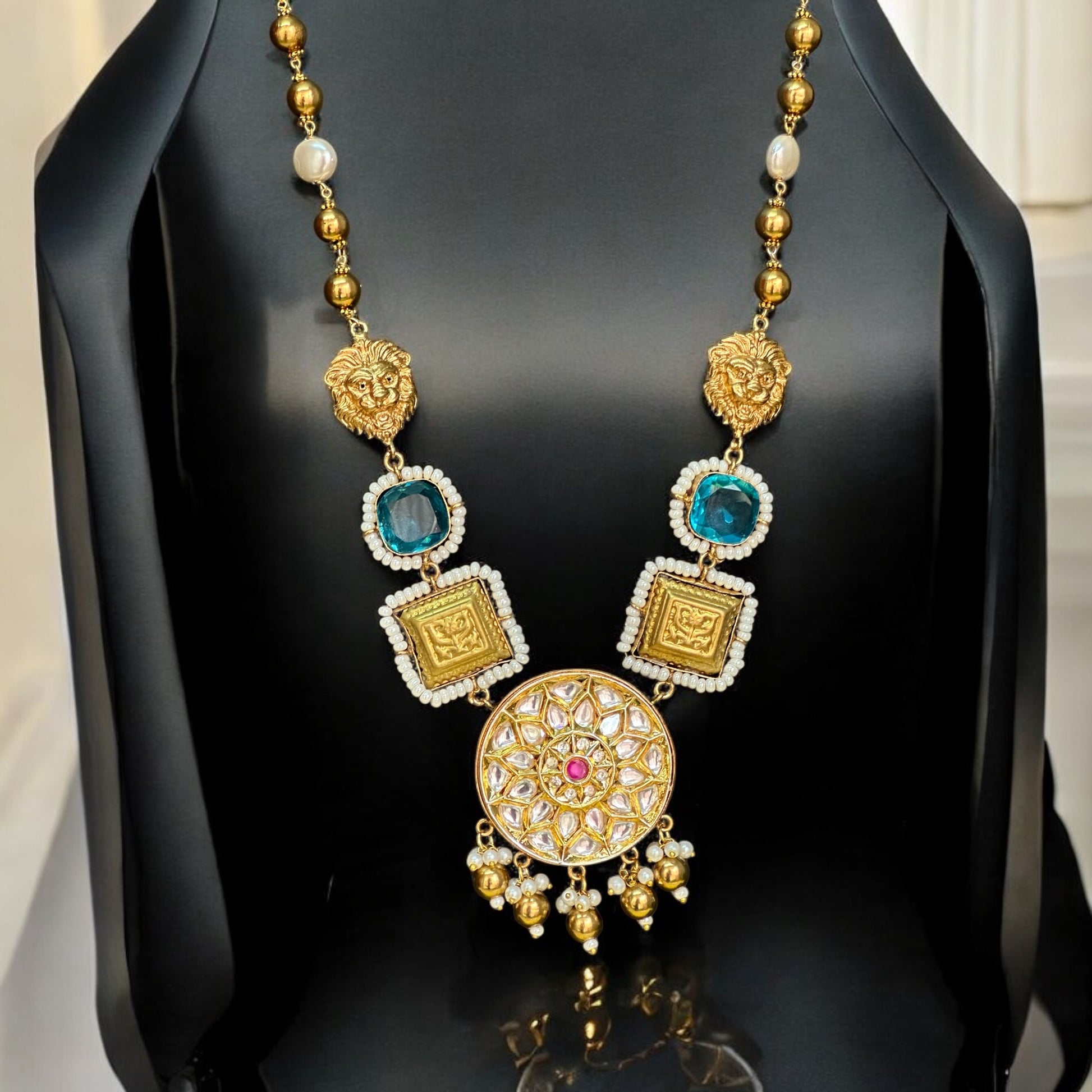 Premium designer Amrapali necklace  featuring gold-toned beads, a lion face, doublet stones in teal and white, a statement circular pendant with intricate detailing and pink gemstone, displayed on a black jewelry bust with an off white background.
