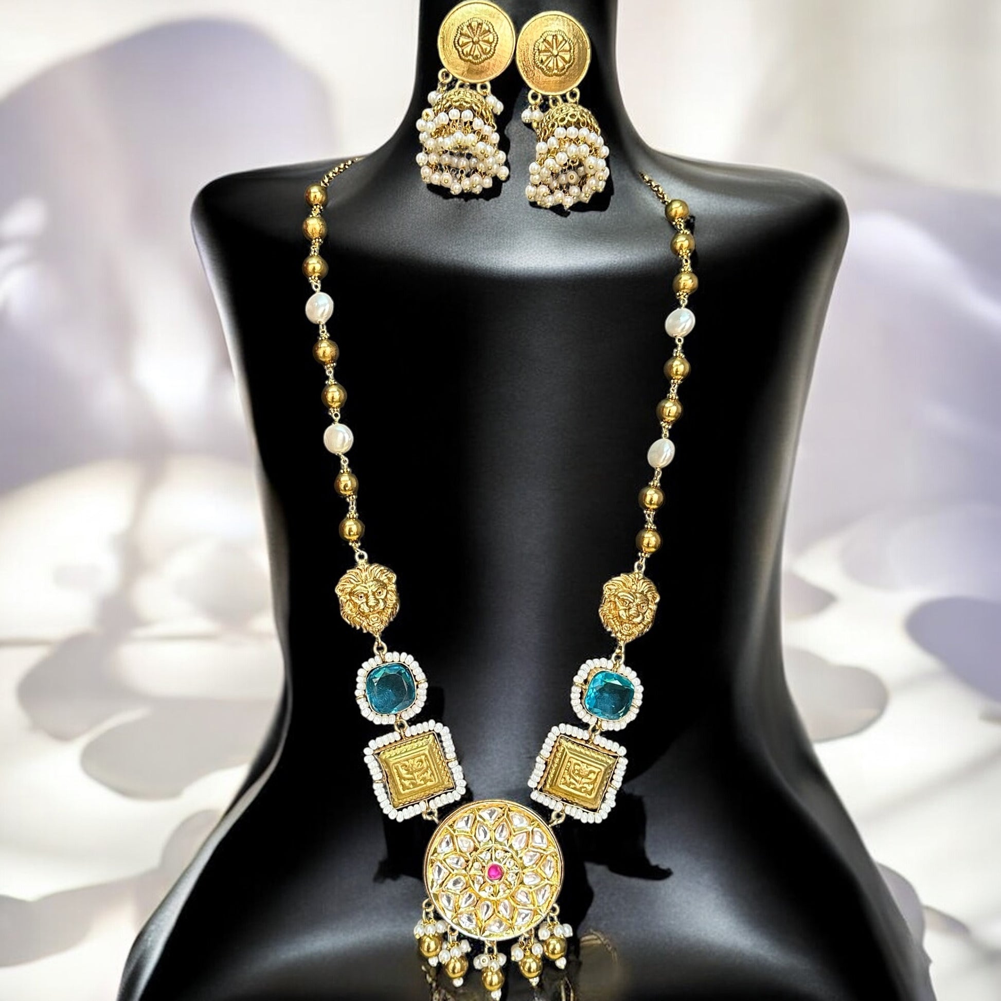 Premium designer Amrapali necklace set featuring gold-toned beads, doublet stones in teal and white, a statement circular pendant with intricate detailing and pink gemstone, paired with pearl drop earrings, displayed on a black jewelry bust with a soft white background.