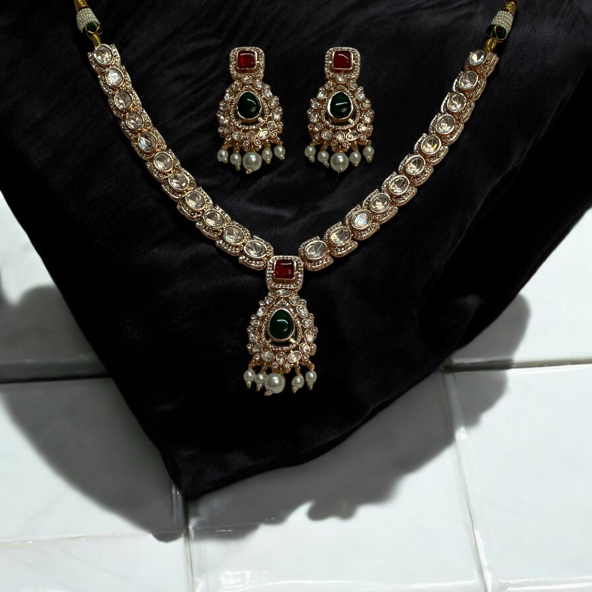 A luxurious Moissanite necklace set with green and red stones, featuring intricate silver foil back finishing, Kundan-style settings, and delicate pearl droplets, displayed on a black bust stand.