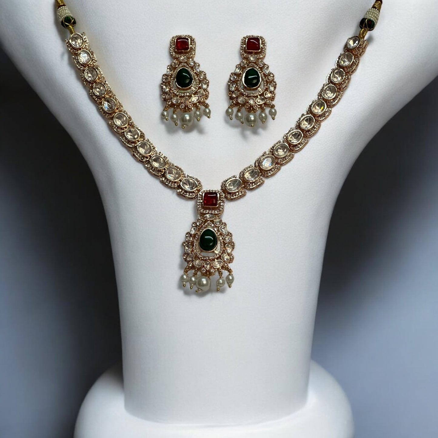 A luxurious Moissanite necklace set with green and red stones, featuring intricate silver foil back finishing, Kundan-style settings, and delicate pearl droplets, displayed on a white bust stand.