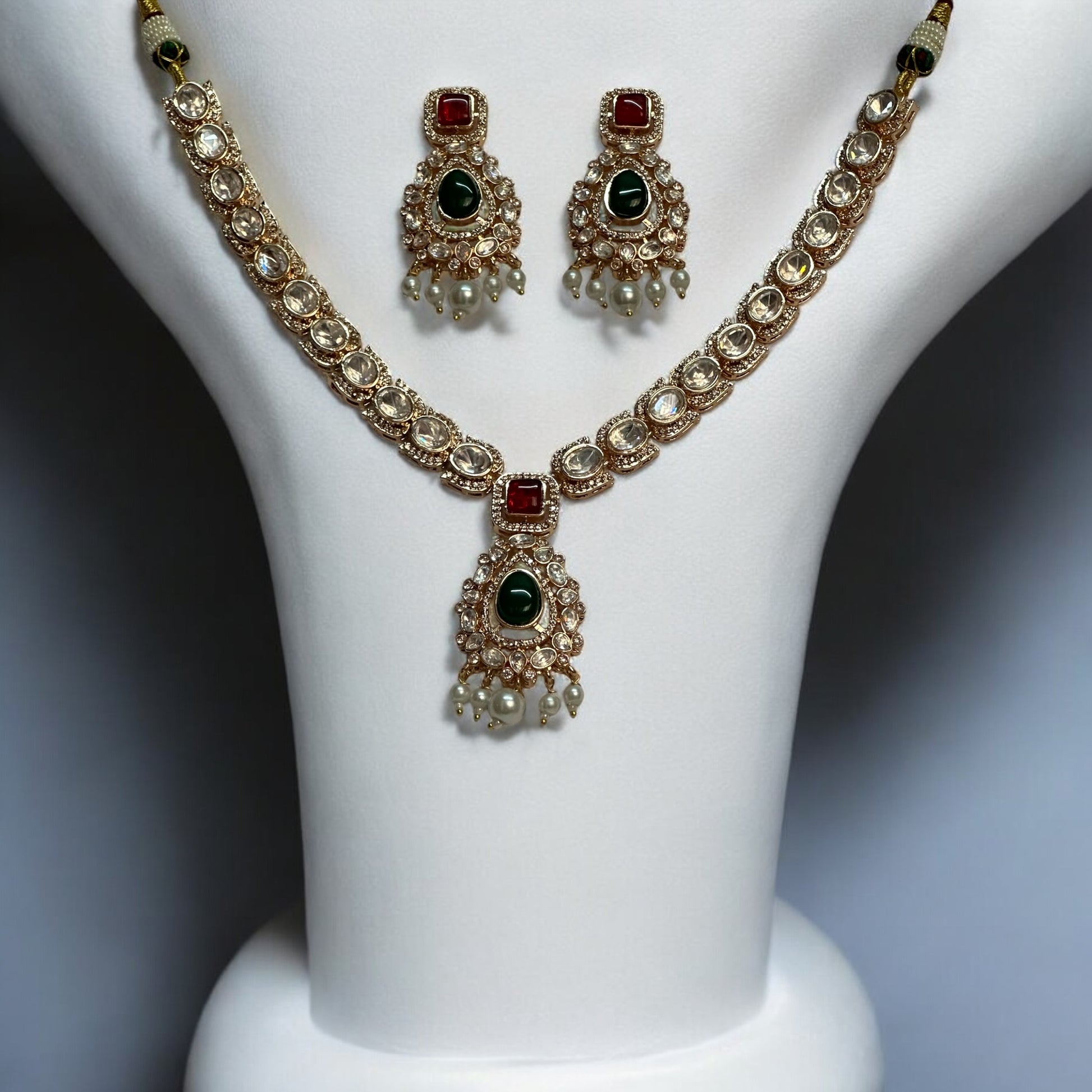 A luxurious Moissanite necklace set with green and red stones, featuring intricate silver foil back finishing, Kundan-style settings, and delicate pearl droplets, displayed on a white bust stand.
