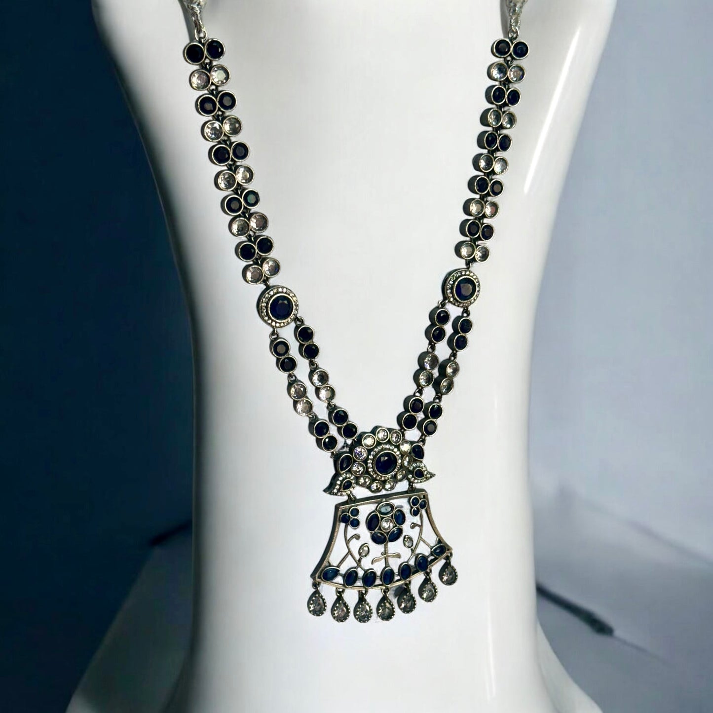 Premium Silver look a like statement necklace in silver and navy blue color stoned placed on a white coloured jewellery display stand.