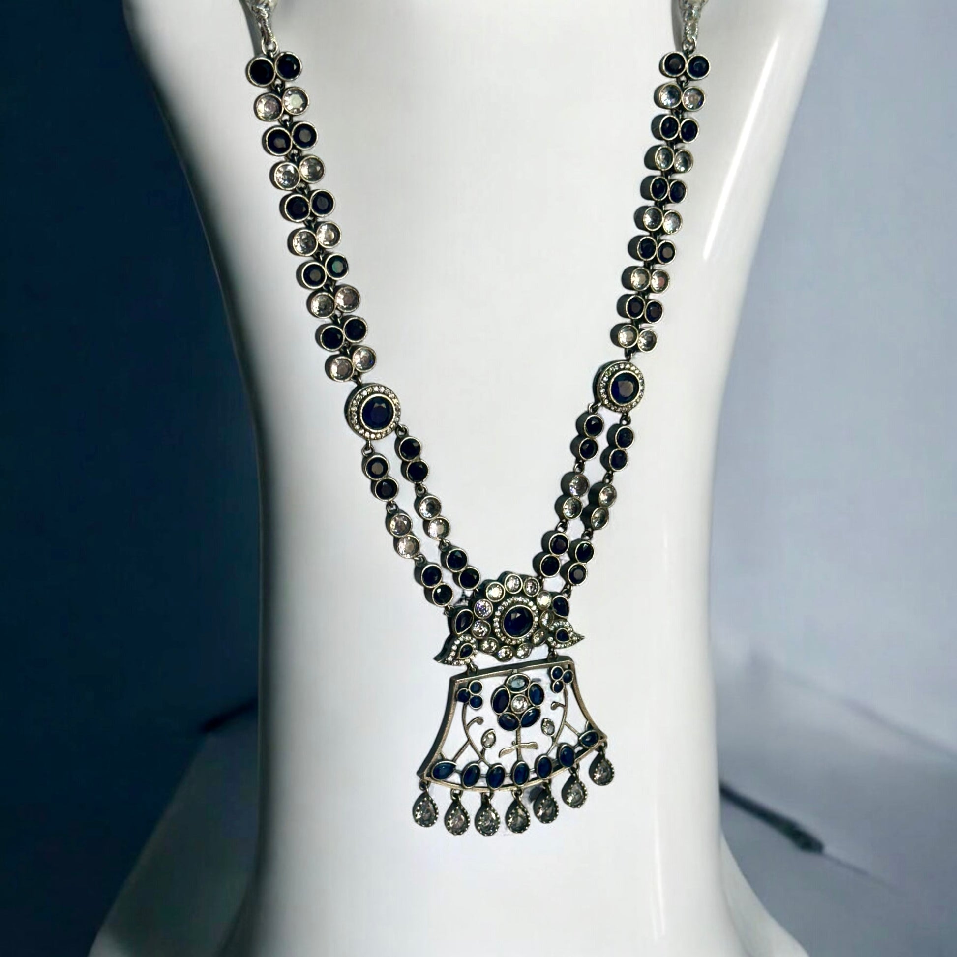 Premium Silver look a like statement necklace in silver and navy blue color stoned placed on a white coloured jewellery display stand.