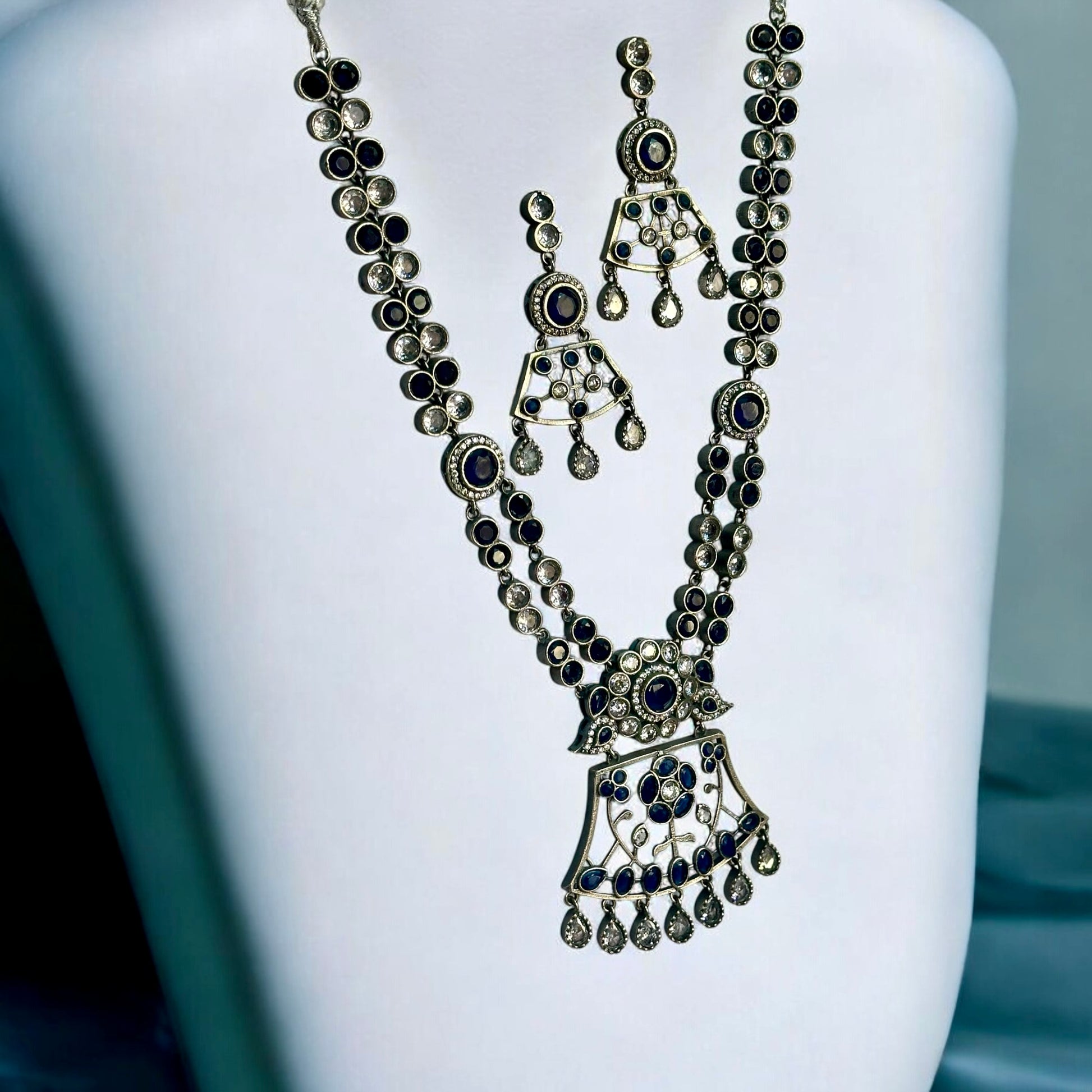 Premium Silver look a like statement necklace and earrings with silver and navy blue color stones placed on a white coloured jewellery display stand.