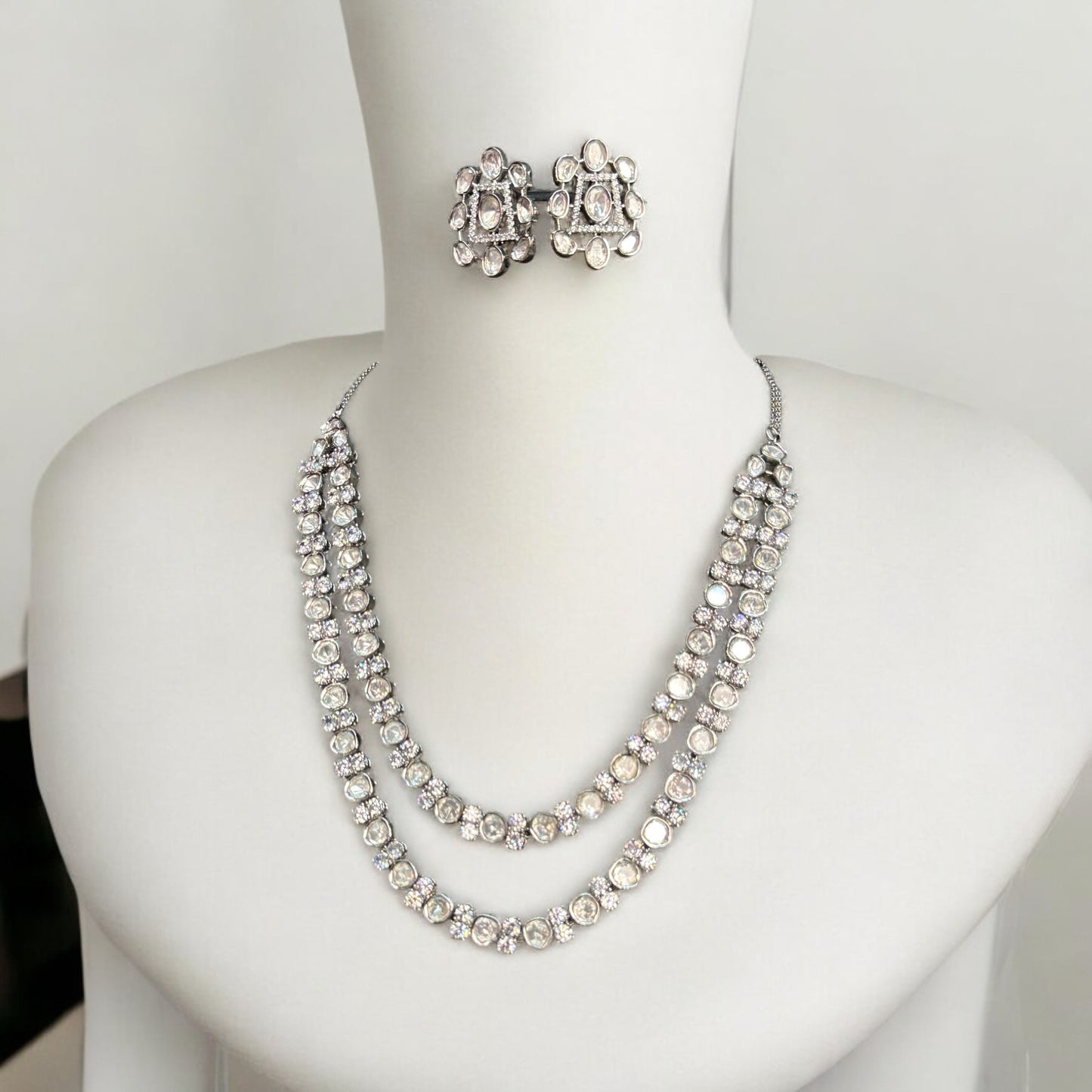 Premium Tyani Kundan double-layer necklace with sparkling stones and matching oversized stud earrings, crafted for luxury occasions. Placed on a white jewellery display stand.