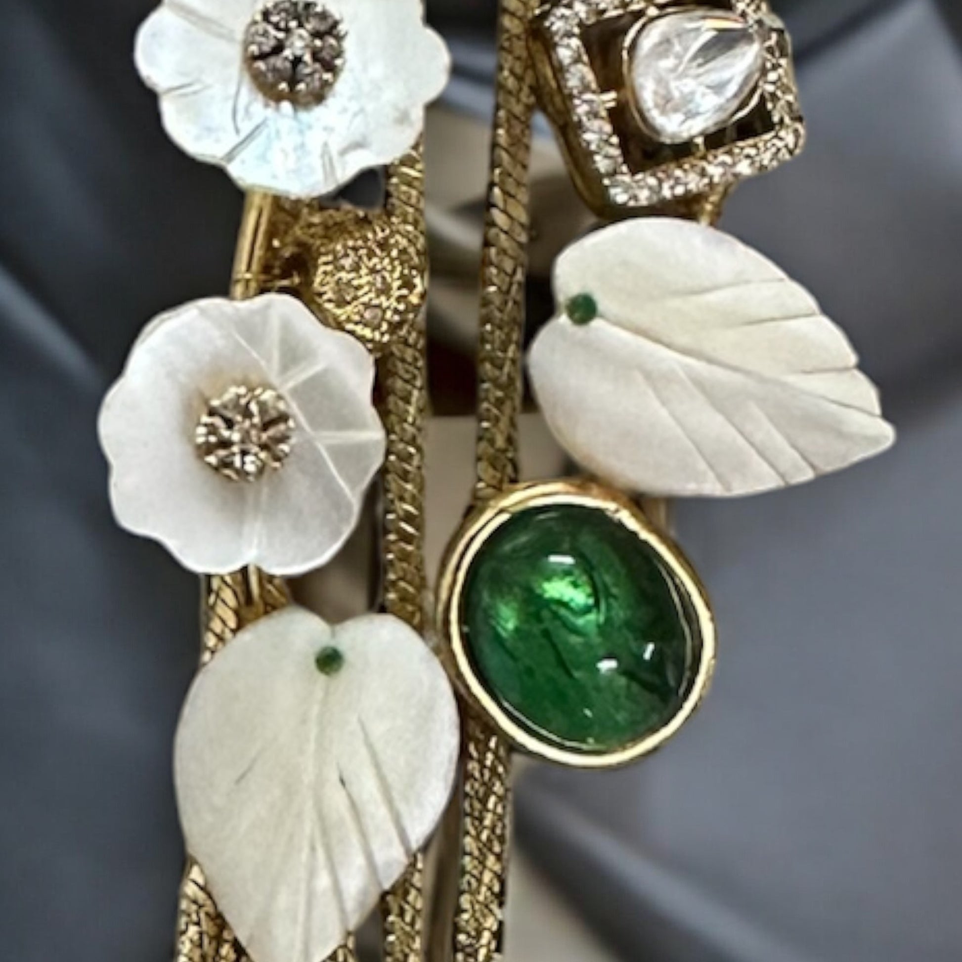 close up of the MOP gold plated moissanite kundan choker design that has white leaves and a green stone with shiny diamond lying on a grey colored fabric.