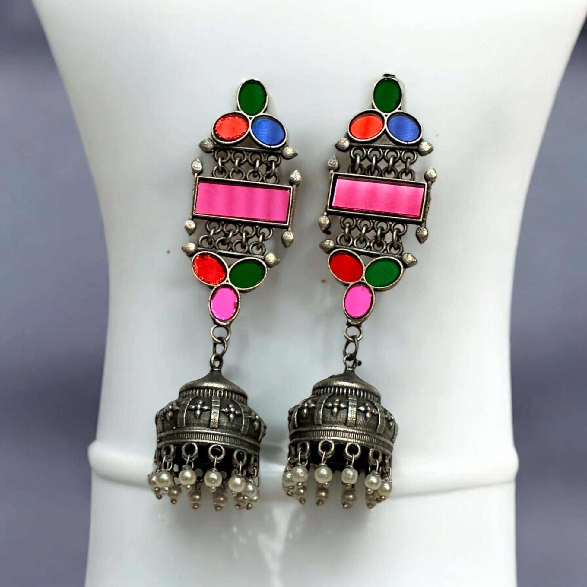German silver earrings with colorful glass inlays in pink, green, blue, and red, featuring a jhumka design with pearl accents placed on a white jewellery display stand.