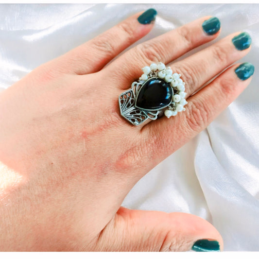 A hand adorned with the Silver Replica Black Ring, showcasing a large teardrop-shaped black stone surrounded by white pearl-like beads and vintage silver detailing, set against a satin background.