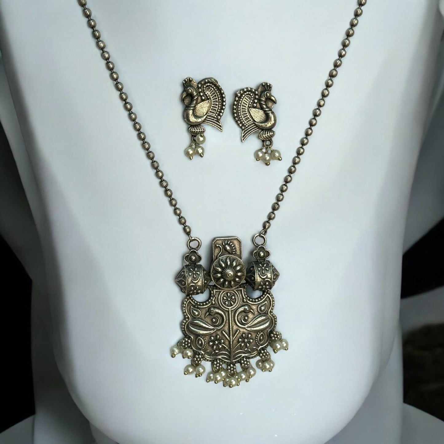 Silver replica designer chain pendant set in antique peacock design with pearl drops and matching earrings displayed on white jewellery stand