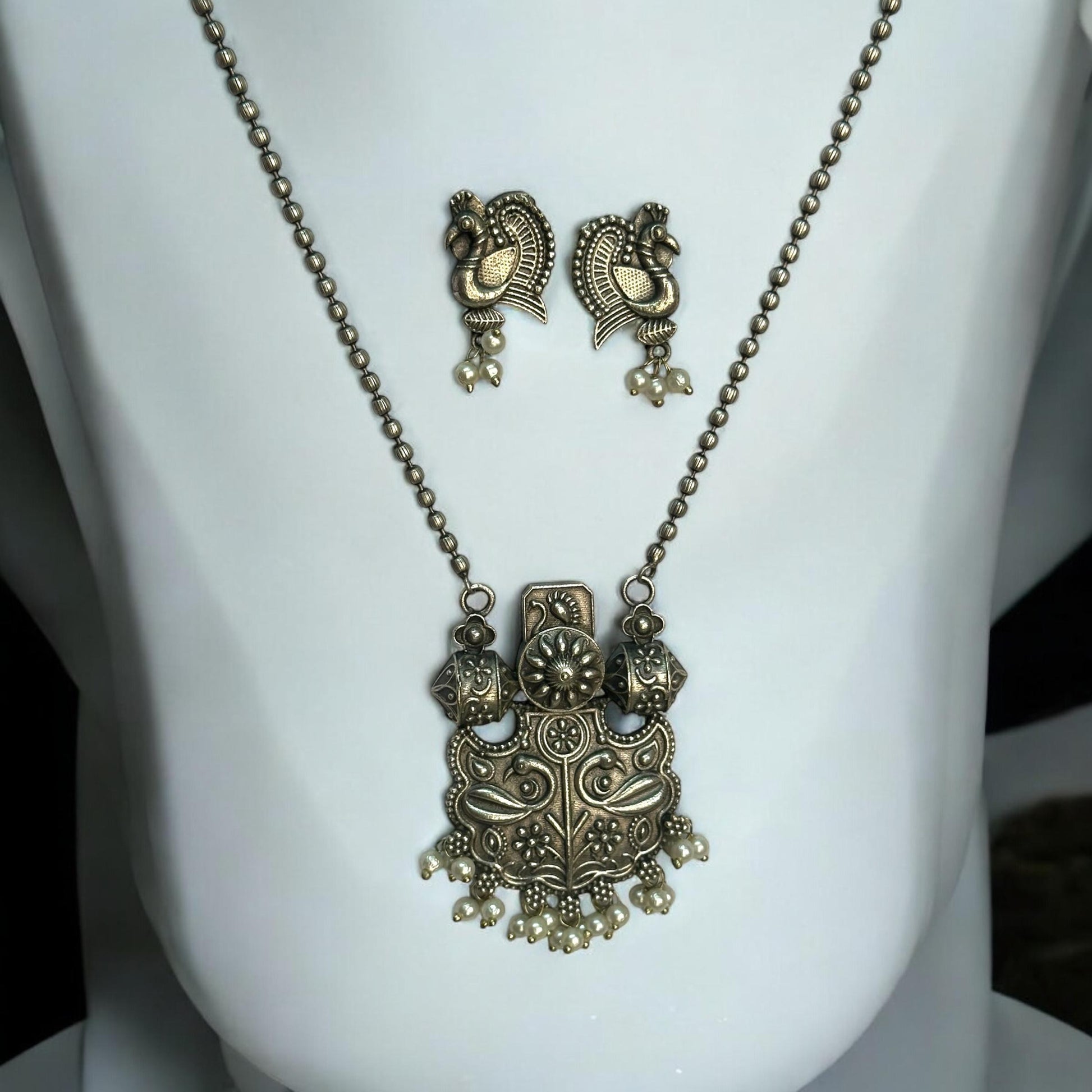 Silver replica designer chain pendant set in antique peacock design with pearl drops and matching earrings displayed on white jewellery stand