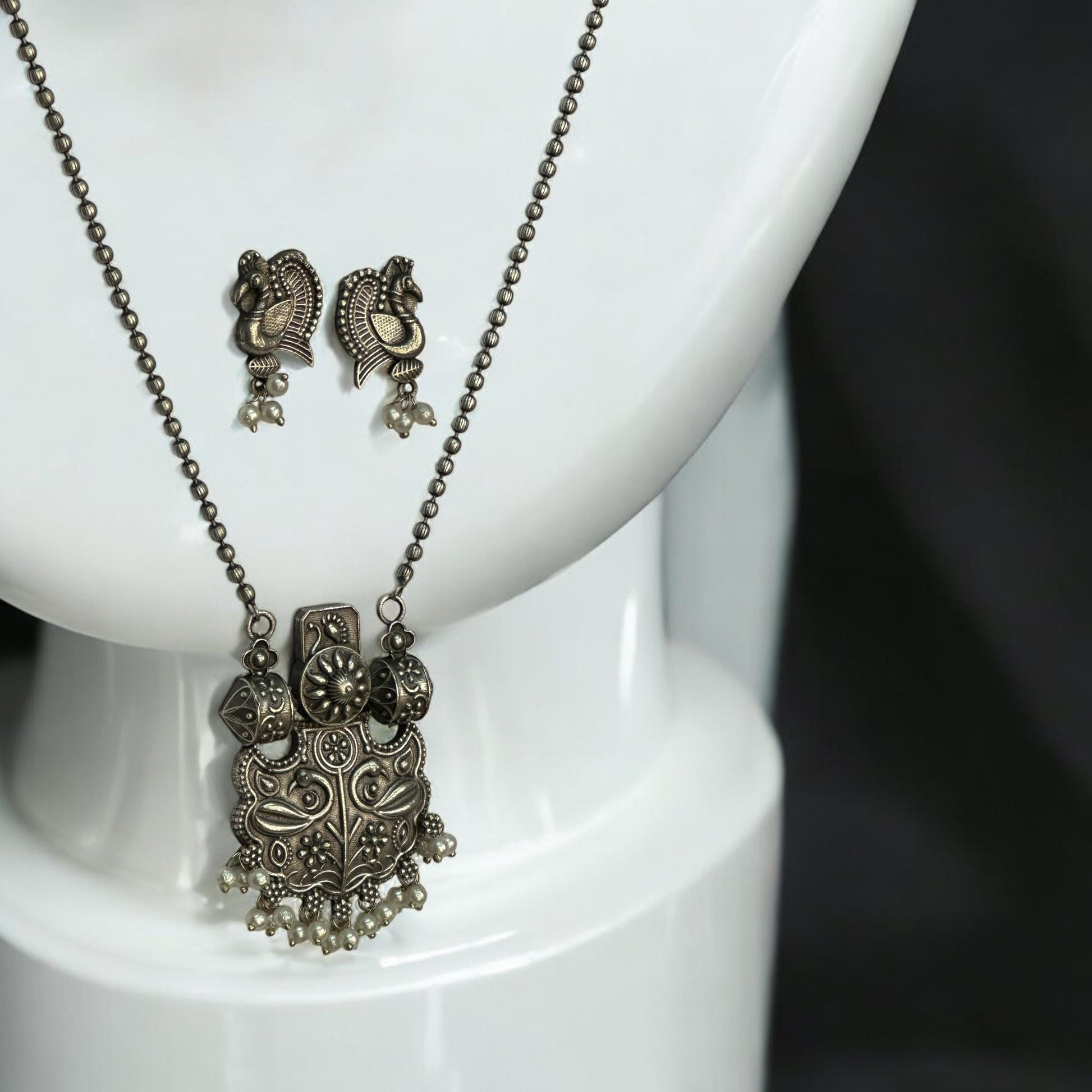 Close look of Silver replica designer chain pendant set in antique peacock design with pearl drops and matching earrings displayed on white jewellery stand
