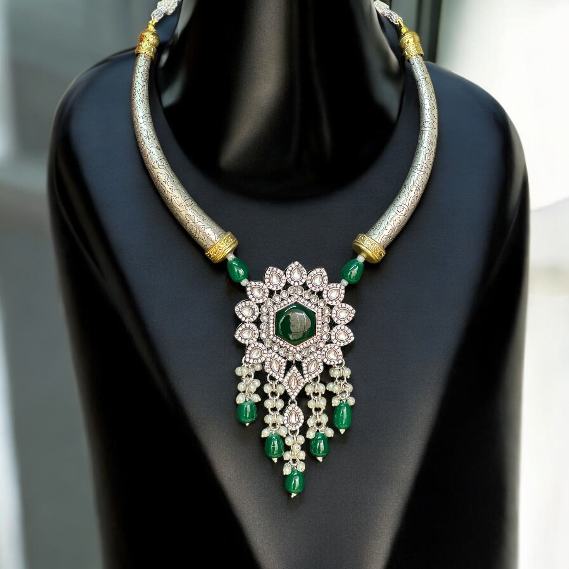 Silver-look alike Hasli necklace adorned with green Monalisa stones and intricate diamond work, featuring a statement floral pendant with pearl and bead danglers, displayed on a black jewelry bust with a soft light background.