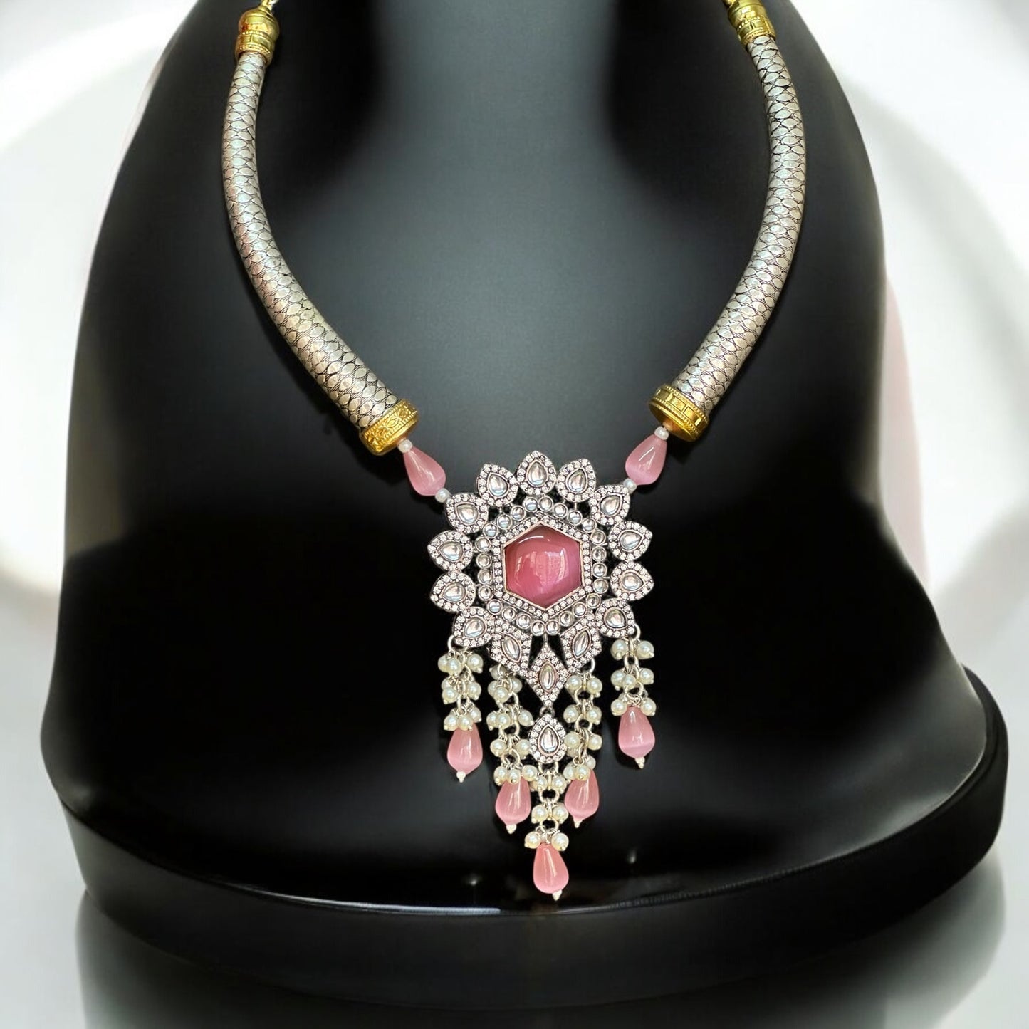 Silver Look-Alike Hasli Necklace adorned with pink Monalisa stones and intricate diamond work. The set is displayed against a black coloured jewellery display stand with a shine and a light white background. 