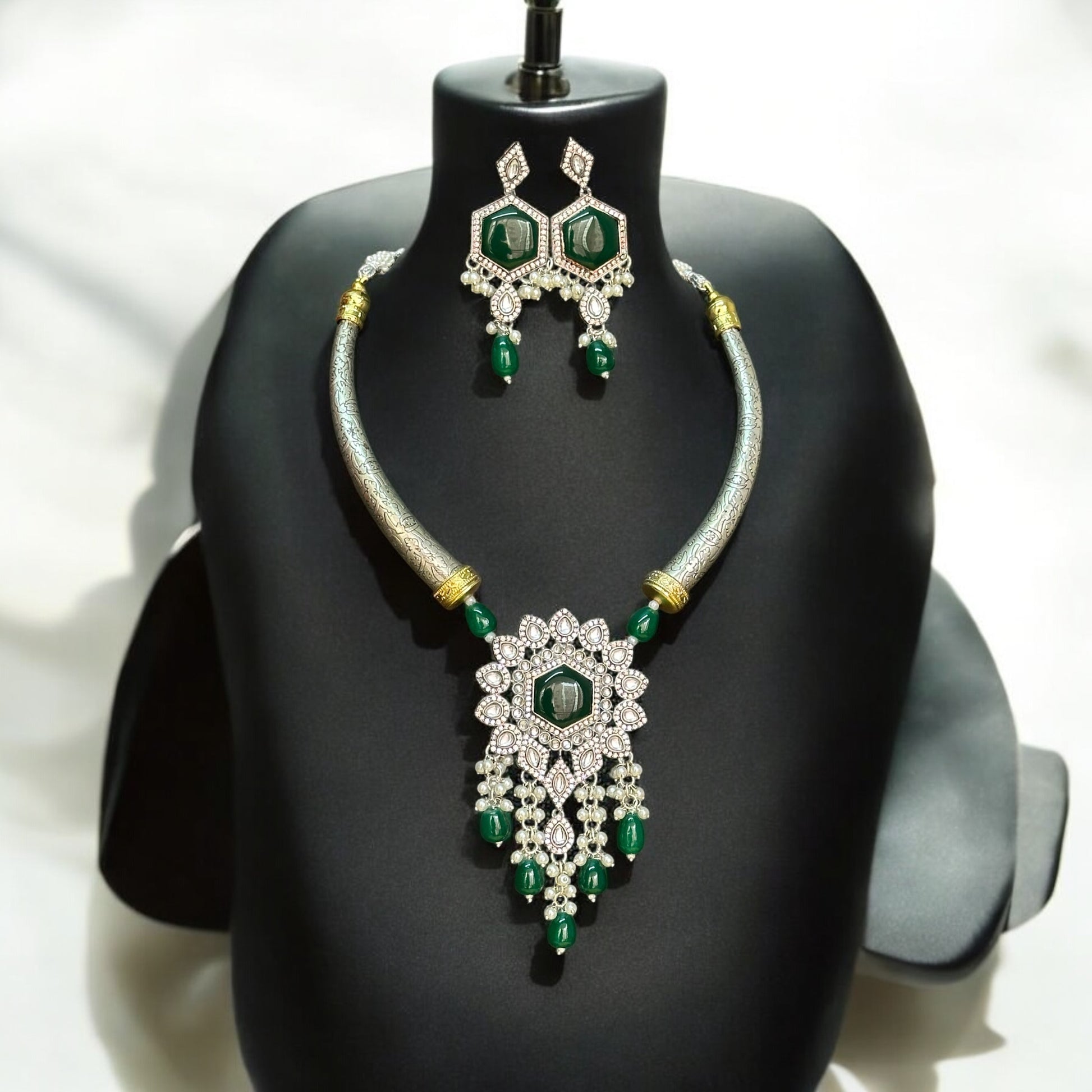 Silver-look alike Hasli necklace adorned with green Monalisa stones and intricate diamond work, featuring a statement floral pendant with pearl and bead danglers, paired with matching green stone earrings, displayed on a black jewelry bust with a soft light background.