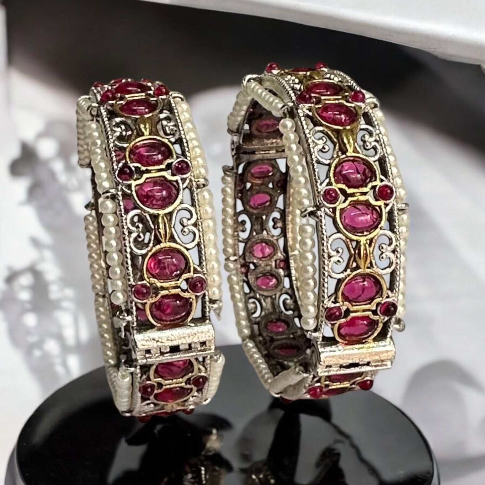 Elegant silver replica bangles with intricate ruby stone detailing and pearl accents placed on a black coloured stand.