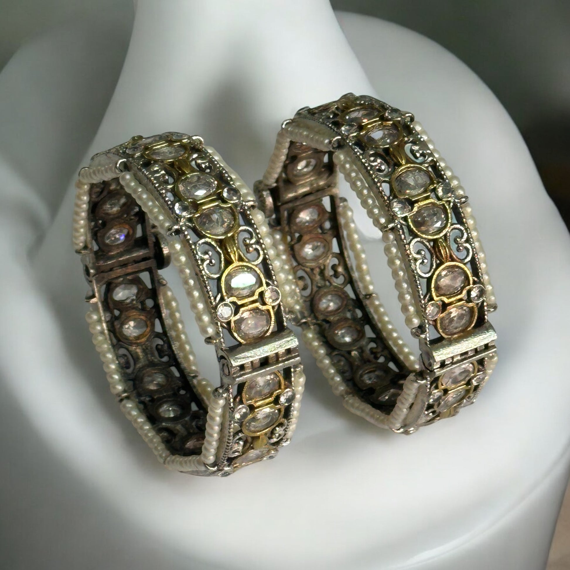 Elegant silver replica bangles adorned with intricate white pearl detailing and sparkling stonework. It is adjustable and is placed on a white jewellery display stand.
