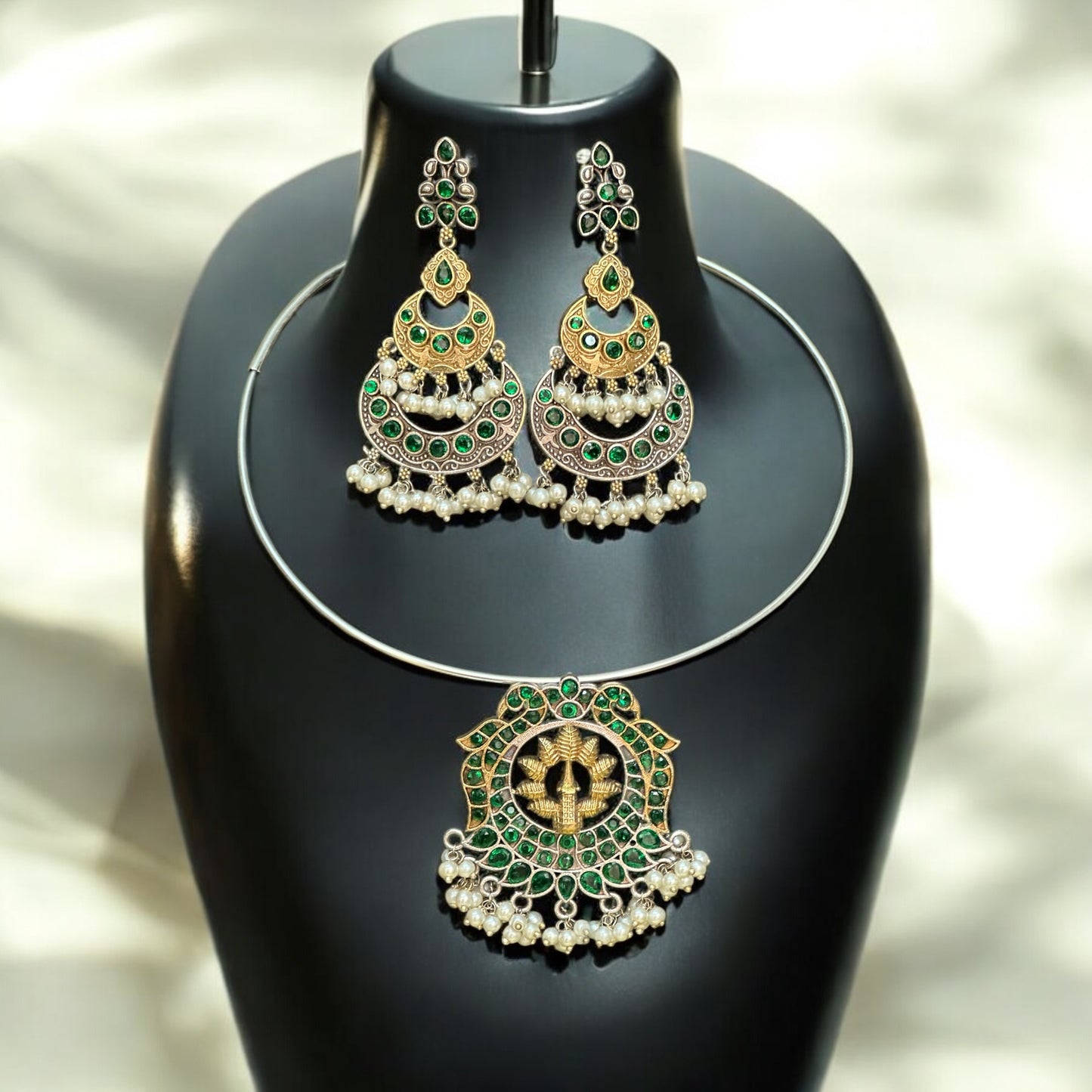 Silver replica Hasli necklace with a round green and gold statement pendant, adorned with pearl danglers, paired with large green and gold jhumka-style earrings featuring intricate detailing, displayed on a black jewelry bust against a soft-lit background.