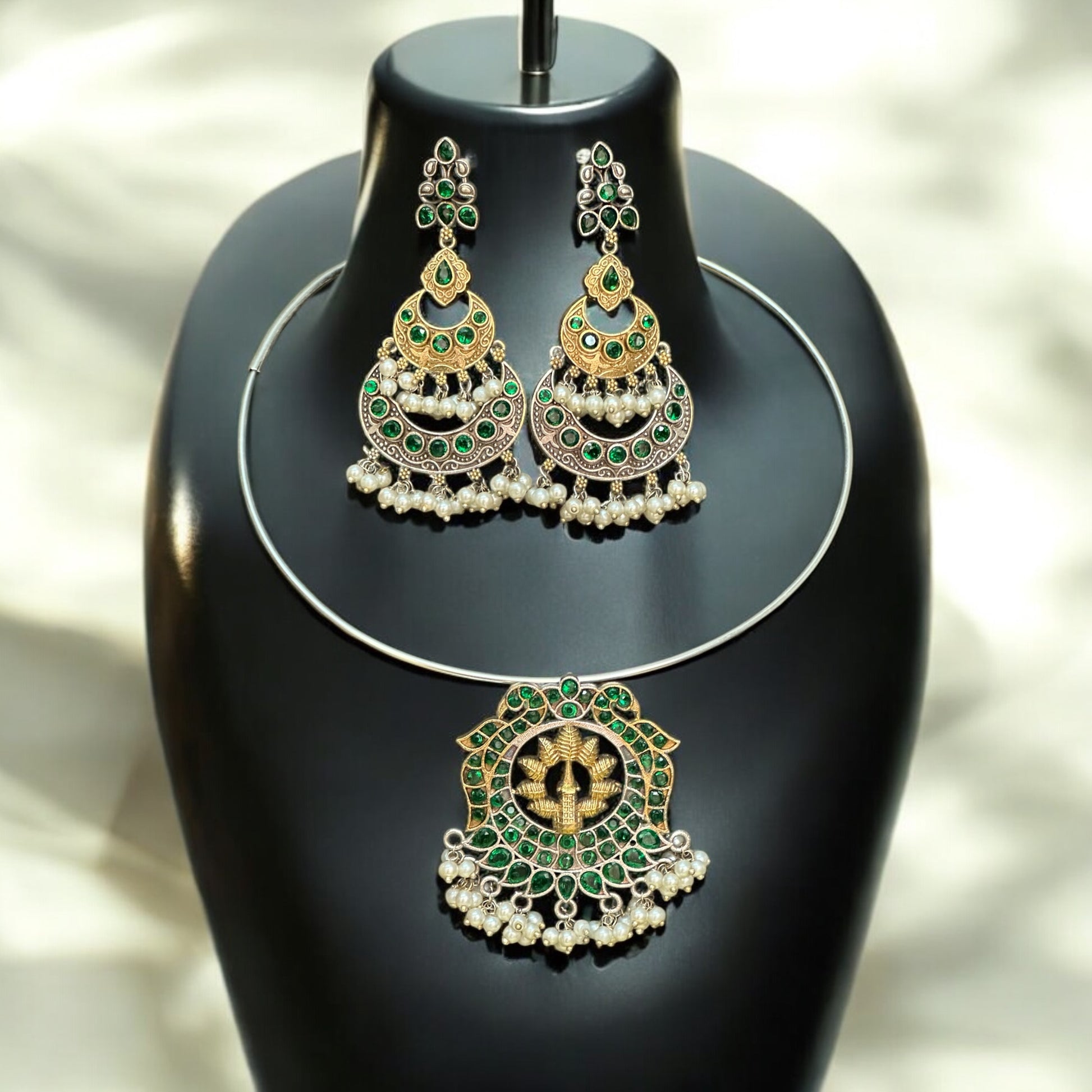 Silver replica Hasli necklace with a round green and gold statement pendant, adorned with pearl danglers, paired with large green and gold jhumka-style earrings featuring intricate detailing, displayed on a black jewelry bust against a soft-lit background.