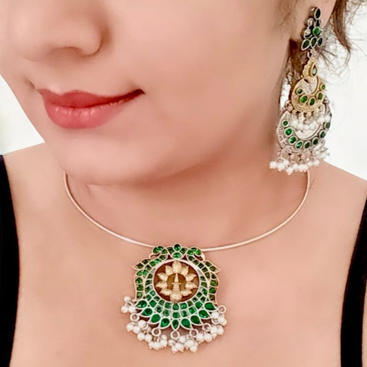 A Model Displaying Silver replica Hasli necklace with a round green and gold statement pendant, adorned with pearl danglers, paired with large green and gold jhumka-style earrings featuring intricate detailing.