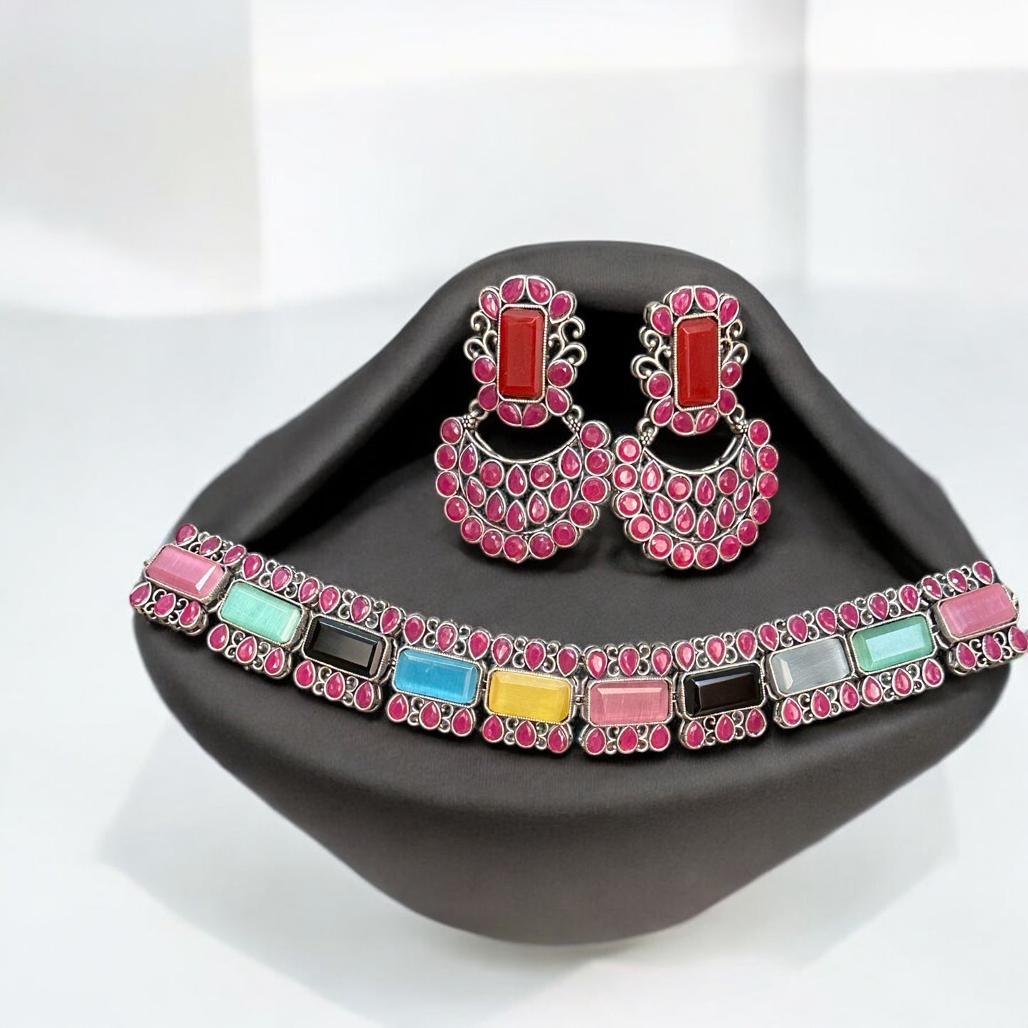 Silver Replica Multicolour Choker Necklace featuring vibrant gemstones in pink, blue, yellow, and more, paired with matching Chandbali-style earrings, displayed on a black velvet stand.