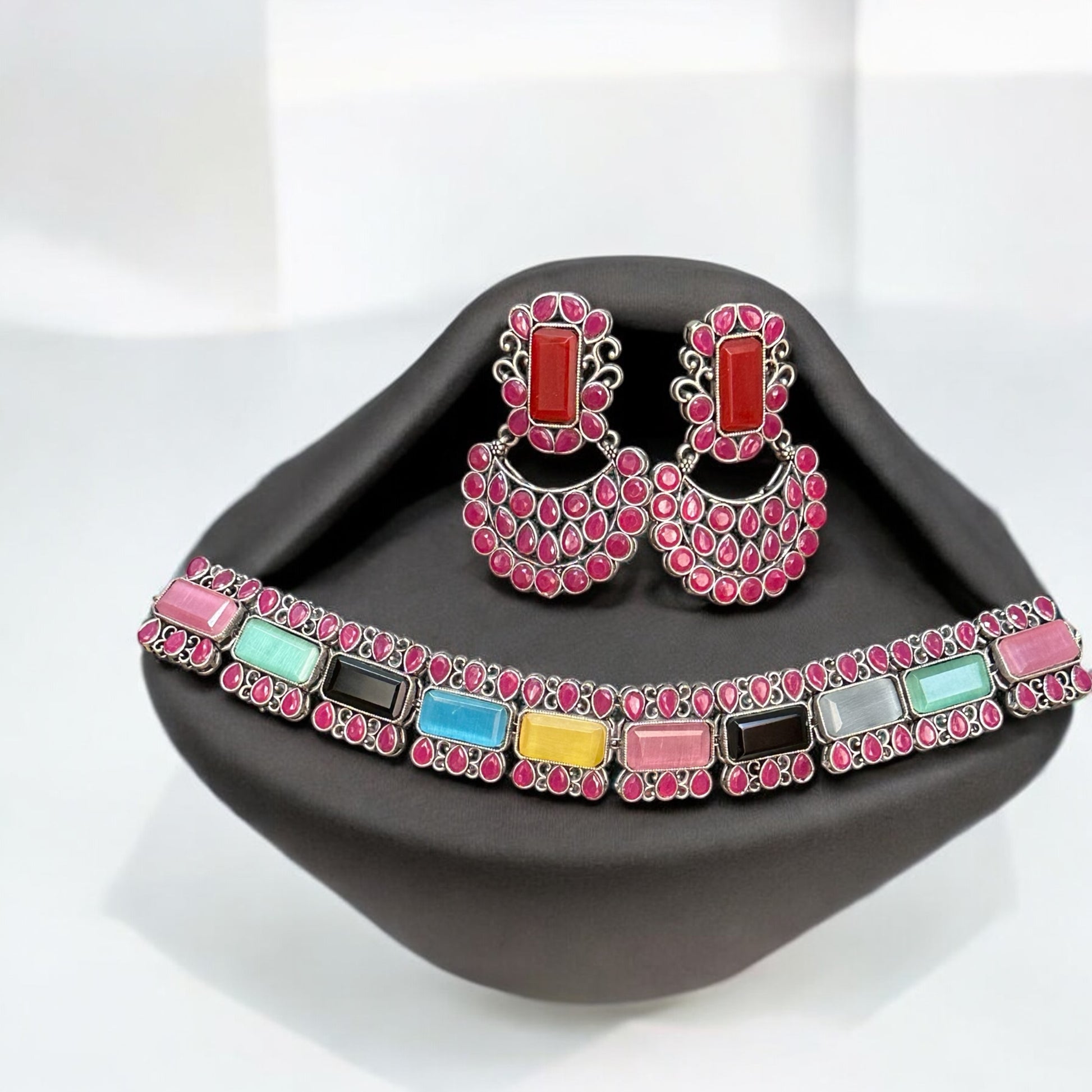 Silver Replica Multicolour Choker Necklace featuring vibrant gemstones in pink, blue, yellow, and more, paired with matching Chandbali-style earrings, displayed on a black velvet stand.