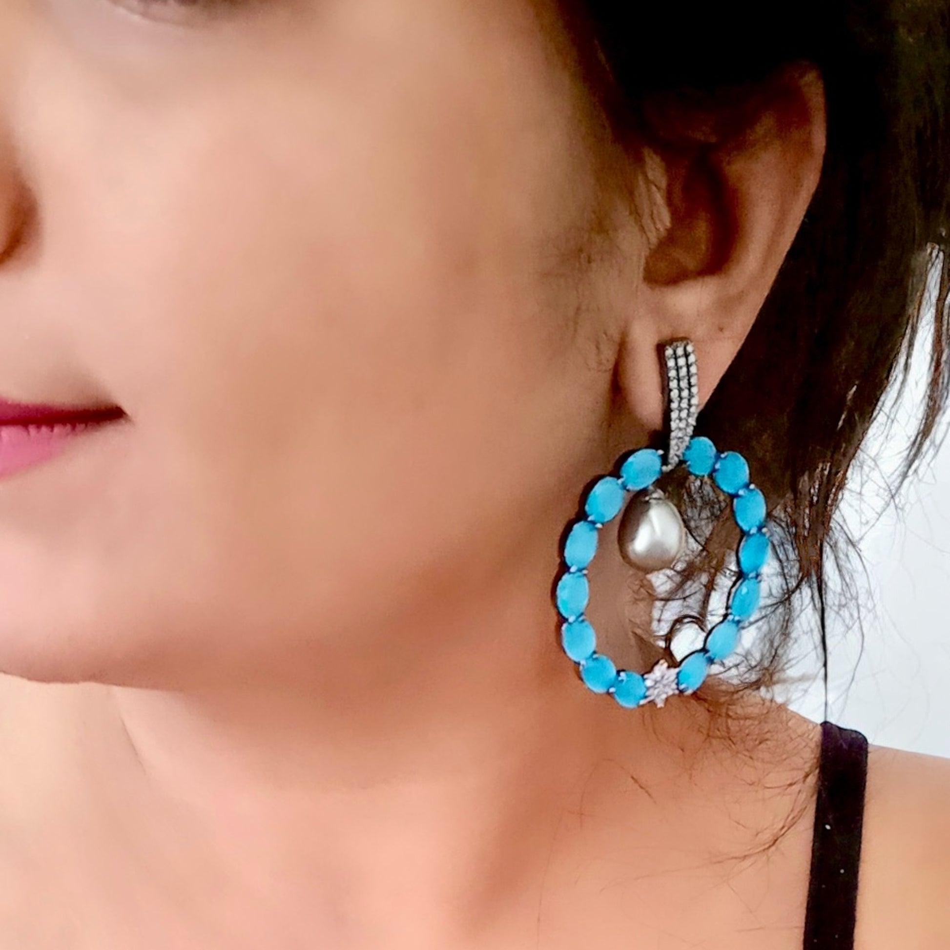 A model wearing and displaying a Statement American diamond earrings featuring a circular blue stone design with a hanging pearl drop, perfect for partywear.