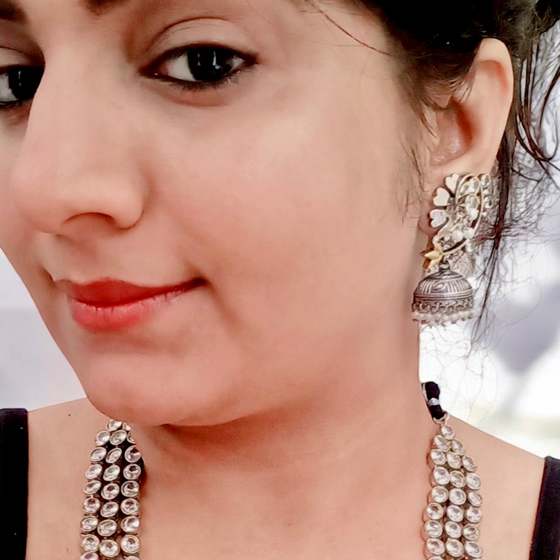 A model displaying a Statement silver replica necklace set featuring three layers of dazzling white stones with matching jhumka earrings, displayed on a black mannequin with intricate detailing.